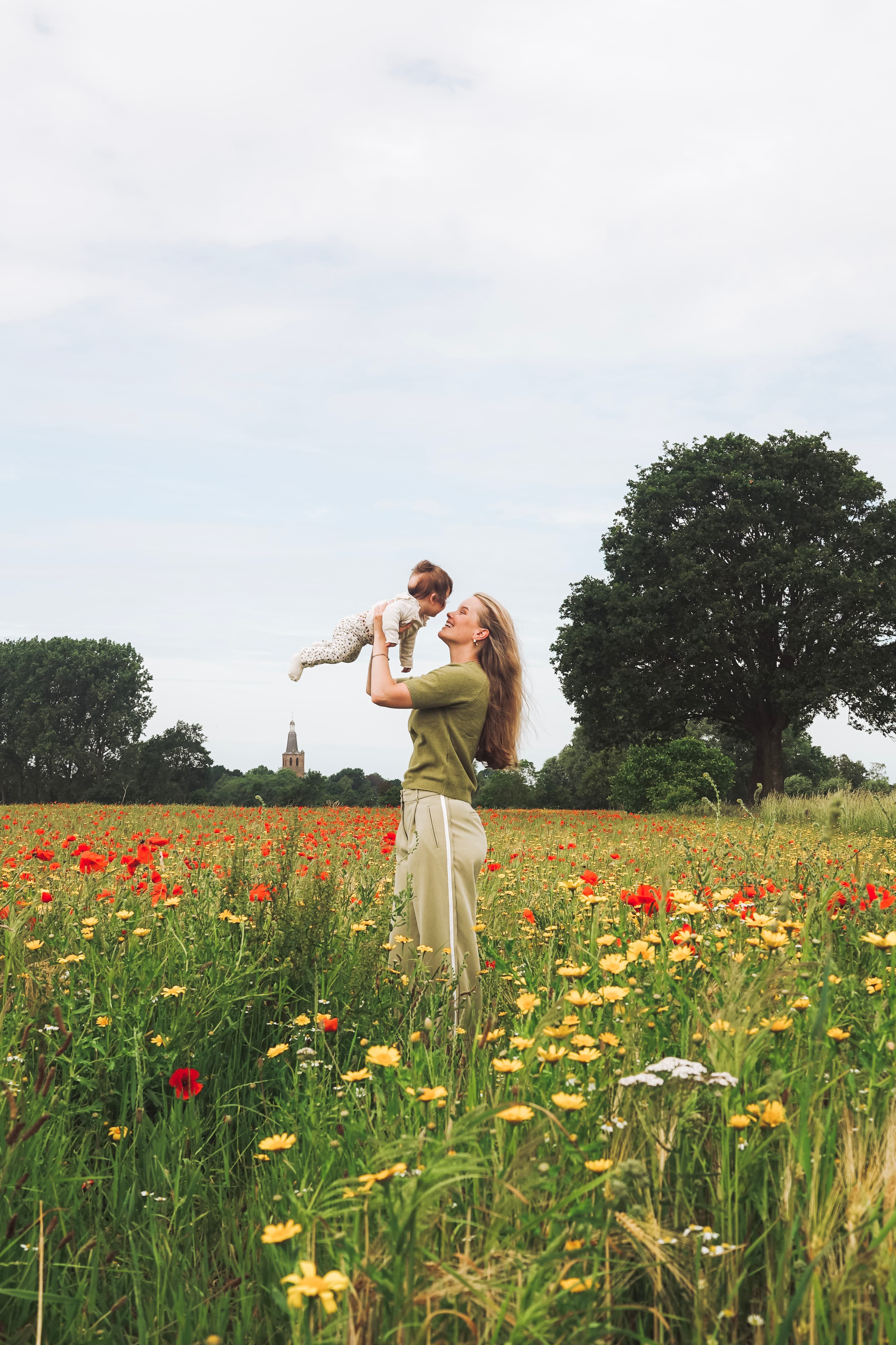 Moeder tilt haar baby lachend omhoog in een veld vol klaprozen en gele bloemen, met de kerktoren van Sint-Oedenrode op de achtergrond